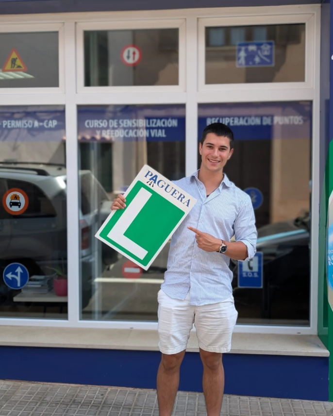 Joven sonriendo y señalando un cartel que dice 'PAGUERA' con una 'L' verde, en la entrada de un centro de formación en conducción, con ventanas que muestran letreros y autos estacionados.