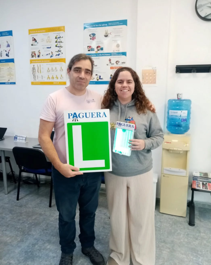 Dos personas, un hombre y una mujer, sosteniendo un cartel de tránsito con una letra 'L' y placas con la palabra 'PAGUERA' en una oficina con posters de salud en la pared y una máquina de agua.