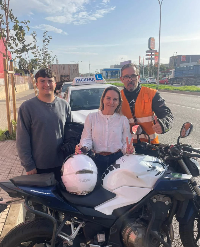 Tres personas posando con una motocicleta en la calle, una mujer en el centro, un hombre con chaleco naranja a la derecha, y un joven a la izquierda, detrás de ellas hay un coche con señal de conductor en formación 'PAGUERA' y un letrero de 'L' azul,