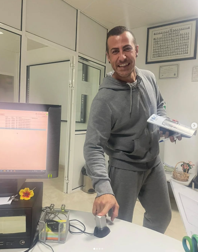 Hombre sonriendo en la oficina, sosteniendo un libro y usando una computadora.