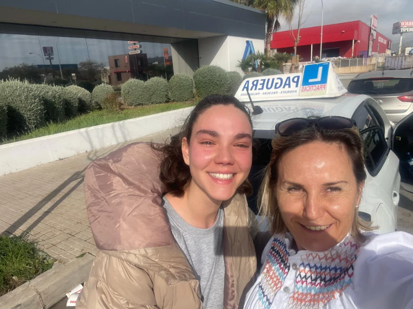 Dos mujeres sonriendo frente a un coche de taxi, con un cartel de licencia de conducir y un edificio en el fondo.