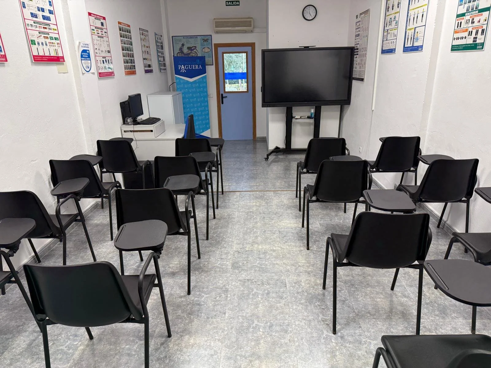 Aula de capacitación con sillas negras con escritorios en estilo pupitre, pantalla grande en la pared, posters educativos en las paredes, computadora y máquina en la esquina, reloj en la pared y puerta con ventana al fondo.