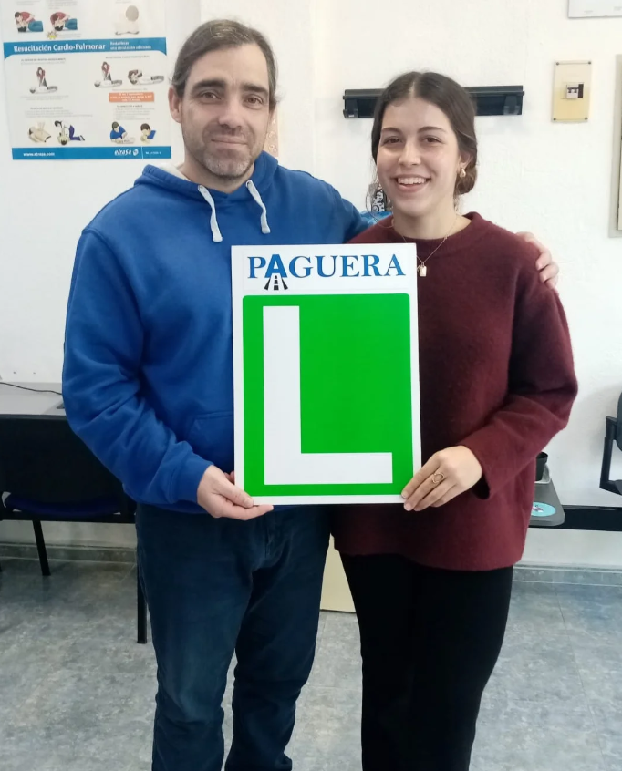 Una pareja sostiene un cartel de aprendizaje de conducir con una letra L grande y la palabra 'PAGUERA' arriba, en un aula de formación vial.