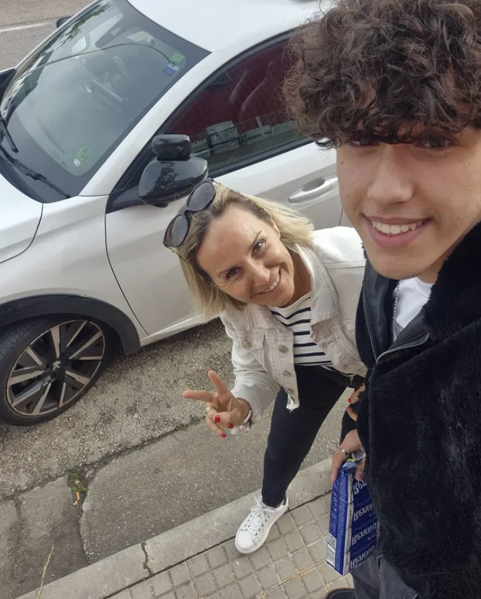 Dos personas sonriendo y haciendo una señal de paz en una calle junto a un automóvil. La mujer lleva gafas en la cabeza y una camiseta a rayas, mientras que el joven sostiene una caja azul.