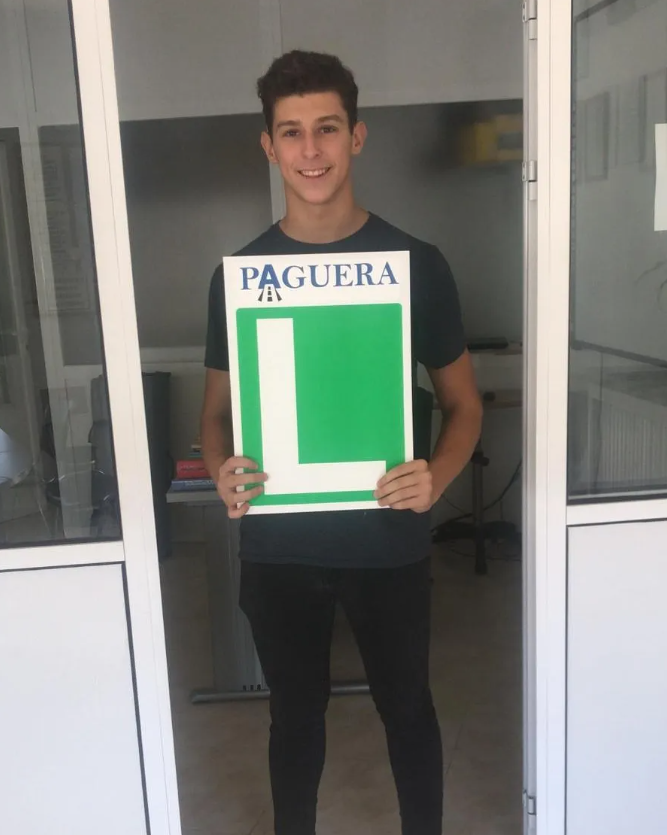 Joven sonriendo sosteniendo un cartel con la letra 'L' en un pasillo.