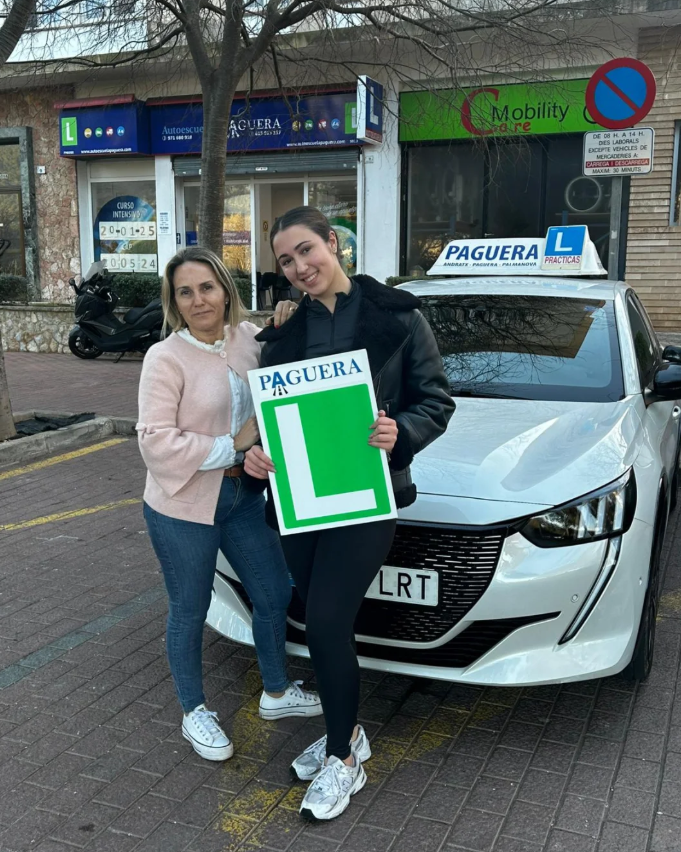 Dos mujeres sosteniendo un cartel de L y un certificado frente a un coche de autoescuela en la calle.