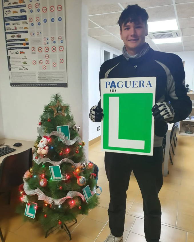 Joven feliz sosteniendo una señal de 'Paguera' junto a un pequeño árbol de Navidad decorado con esferas y luces en una oficina.