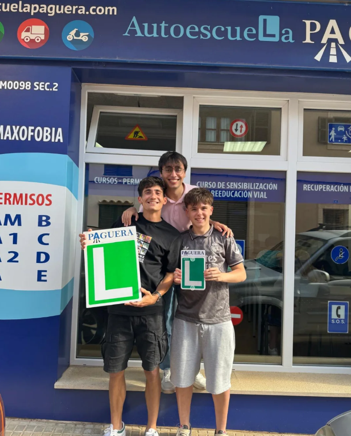 Tres jóvenes con placas de 'L' de conductor en entrenamiento, sonriendo frente a una academia de manejo.