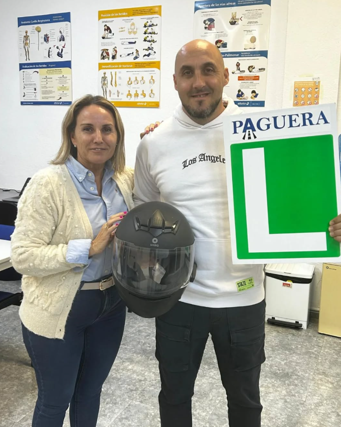 Una mujer y un hombre posan en una sala de clases. La mujer sostiene un casco de motocicleta y el hombre muestra un cartel con una letra 'L' y la palabra 'PAGUERA'. Detrás hay carteles con información sobre primeros auxilios y anatomía, y en la esqui