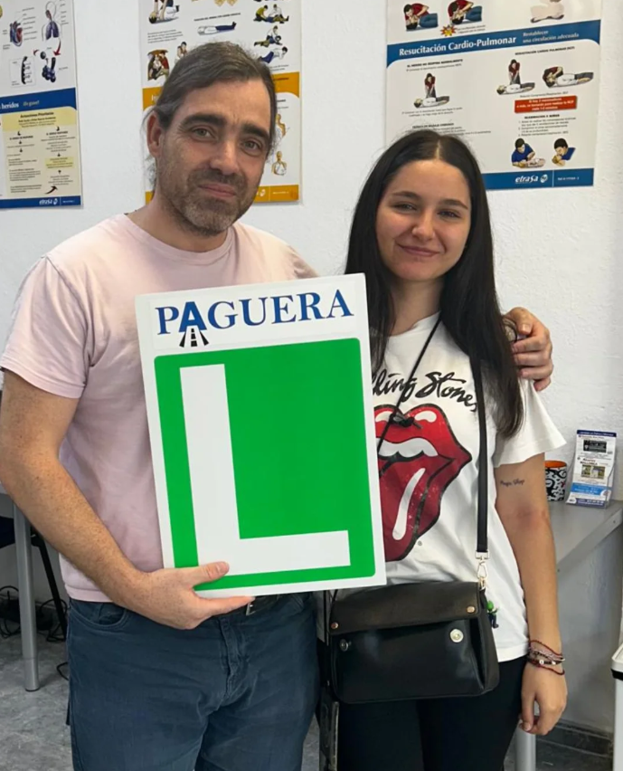 Hombre y mujer en una clínica de primeros auxilios, con carteles de reanimación en el fondo, sosteniendo un cartel grande con una letra L en color verde y la palabra "Paguera" en la parte superior.