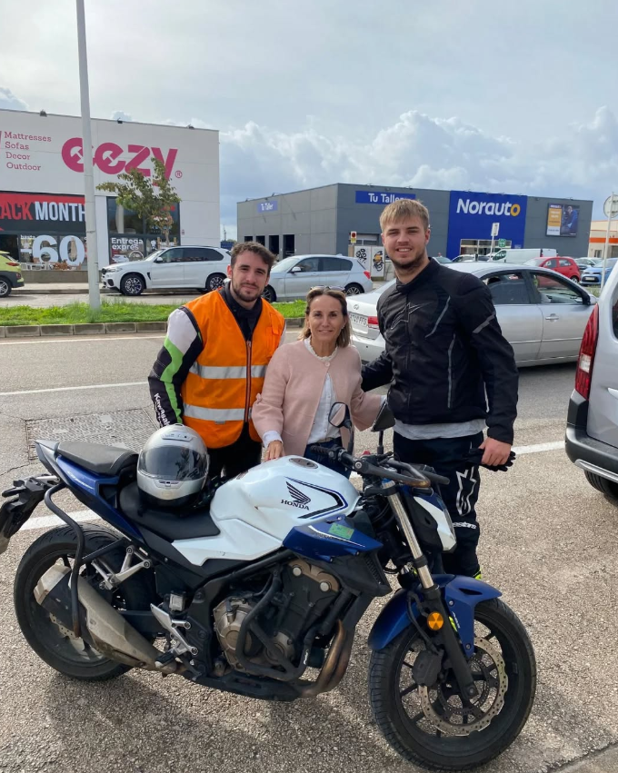 Tres personas posando junto a una motocicleta Honda en un estacionamiento, en un día nublado, con establecimientos comerciales de fondo.