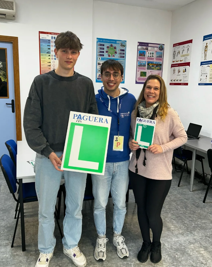 Tres personas en una sala, sosteniendo señales de aprendizaje de conducir, la de la izquierda con una señal de principiantes 'L', el de en medio con una gorra y una chaqueta azul y el de la derecha una señal de 'P' para principiante, todos sonriendo.