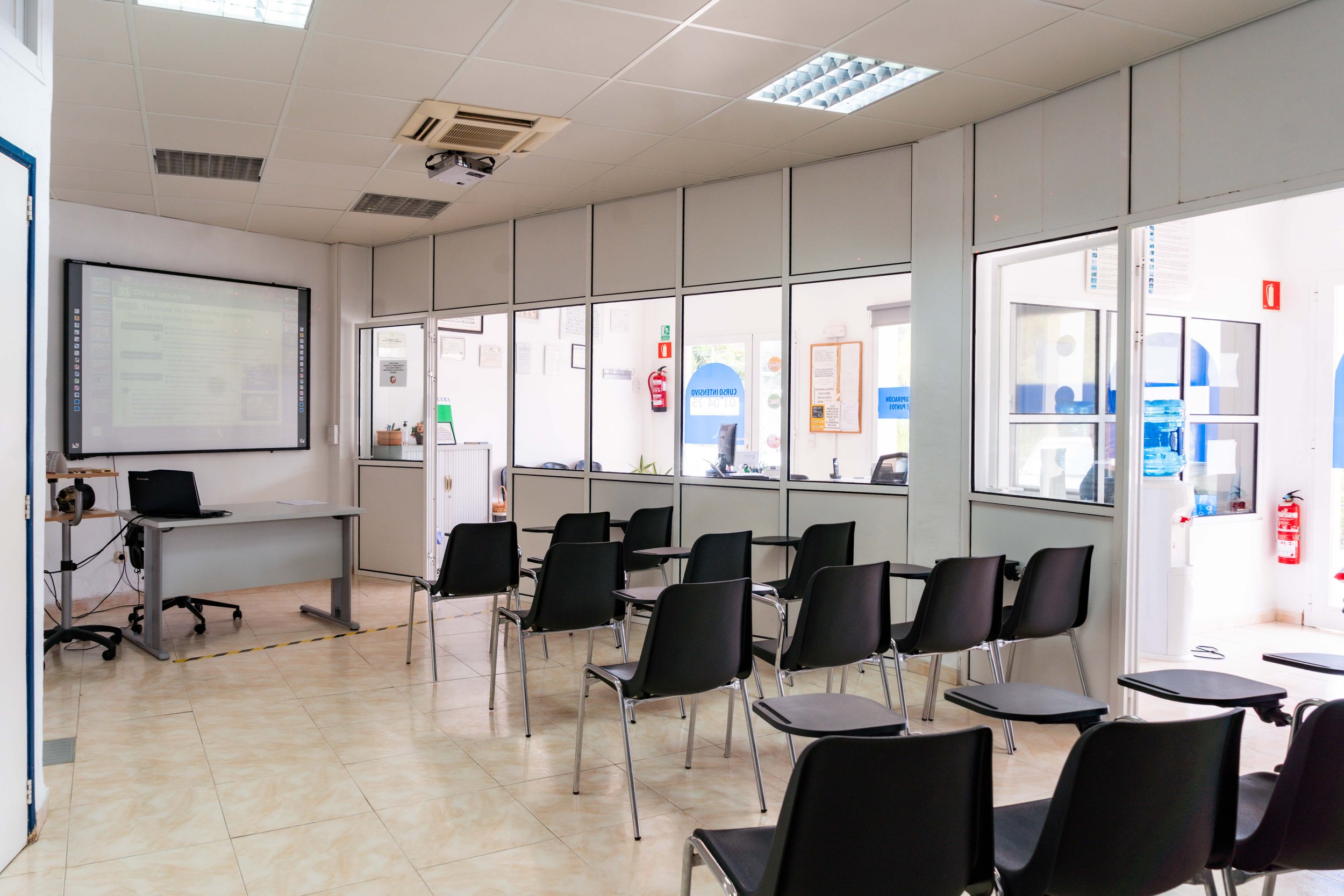 Sala de espera con varias sillas negras, una pizarra digital en la pared, ventanas grandes, un escritorio con una computadora y una puerta de entrada con ventanas de vidrio que muestran un área de oficina, con señalización y extintor.