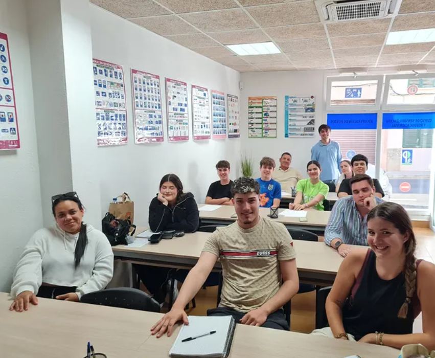 Grupo de estudiantes en un aula durante una clase o reunión, con una mujer y un hombre en el fondo y varias cartelones en la pared.