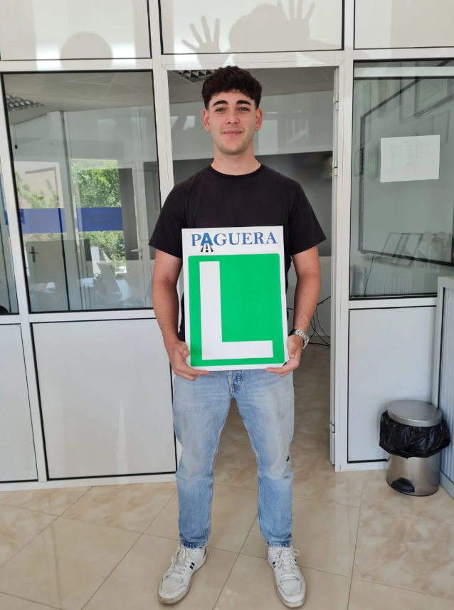 Joven con camiseta negra sostiene un cartel con la letra 'L' verde y la palabra 'PAGUERA' en la parte superior, en un ambiente interior con ventanas.