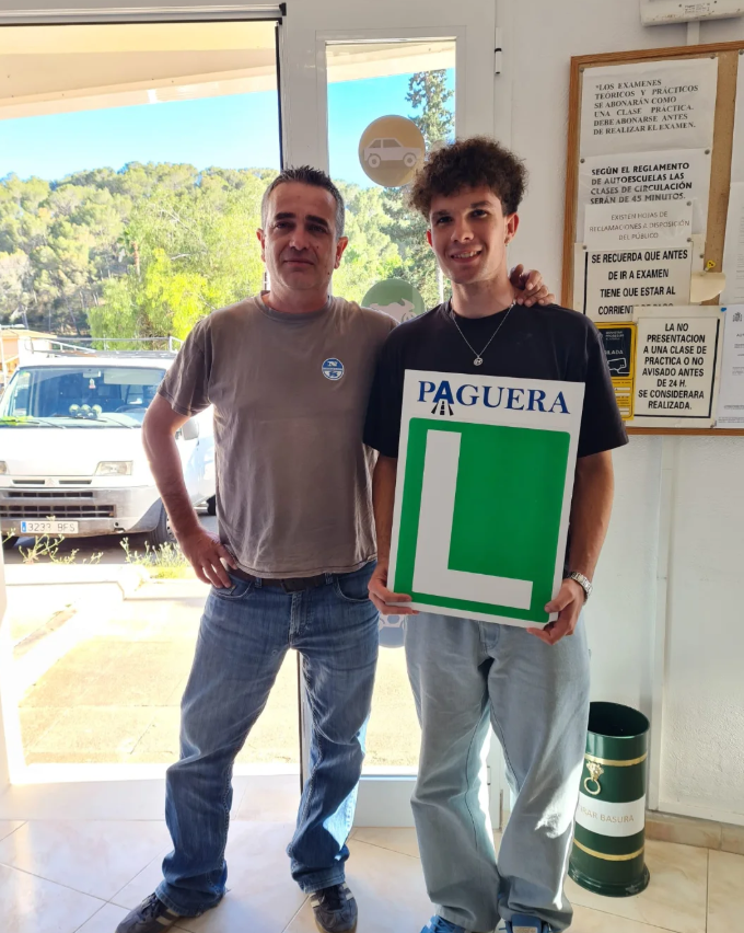 Dos personas, un hombre mayor y un joven, posan en un interior con una puerta de vidrio que muestra vegetación y vehículos afuera. El joven sostiene un cartel de aprendizaje de manejo con una letra 'L' grande y la palabra 'PAGUERA' en la parte superi