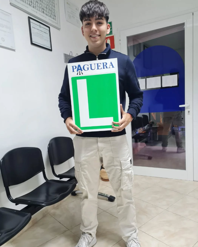 Joven sonriendo sosteniendo un cartel con una 'L' grande y la palabra 'PAGUERA' en la parte superior, en una sala con sillas negras.