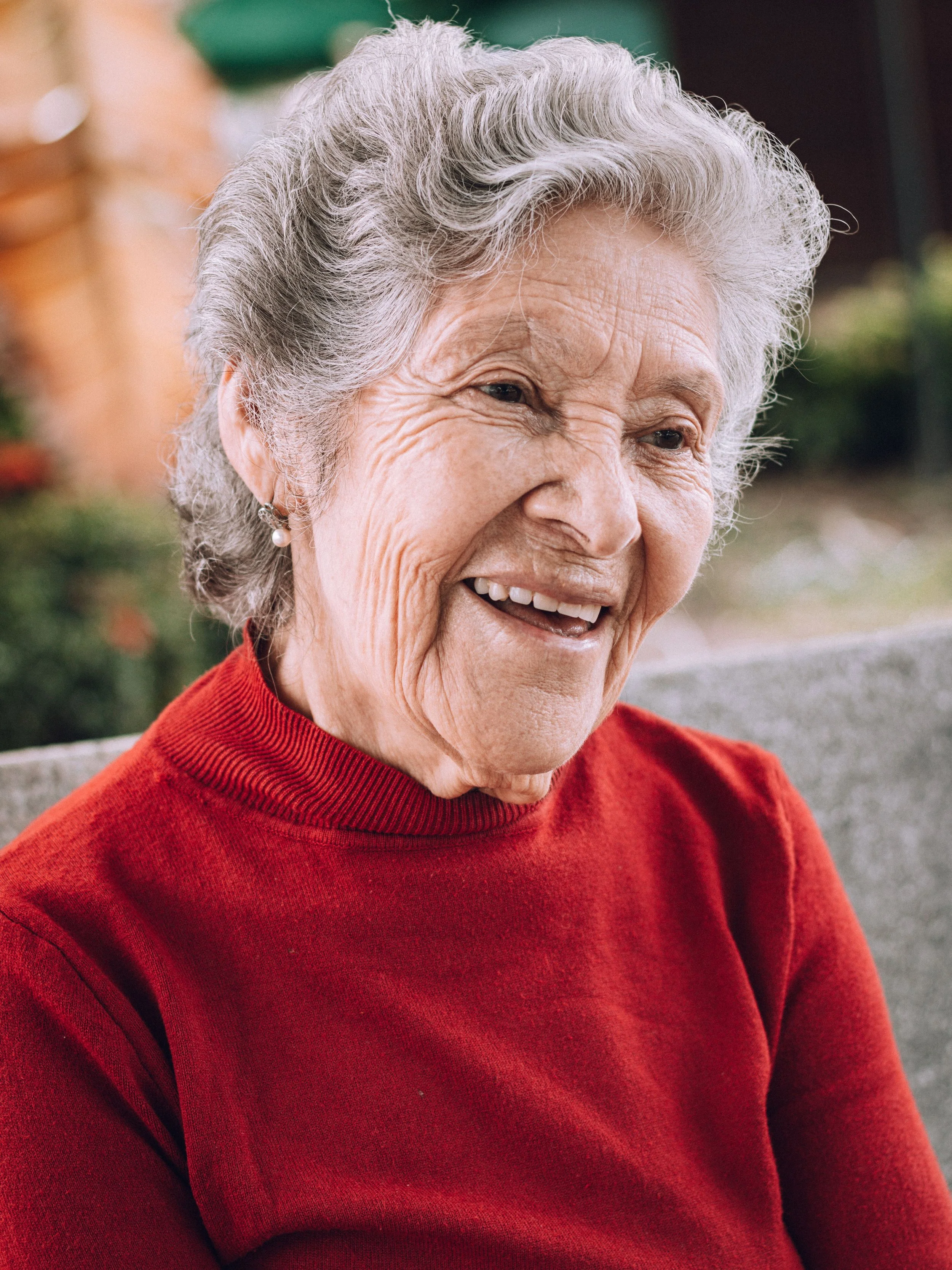 Close-up of an elderly woman with gray hair smiling, wearing a red sweater and pearl earrings, outdoors with blurred background.