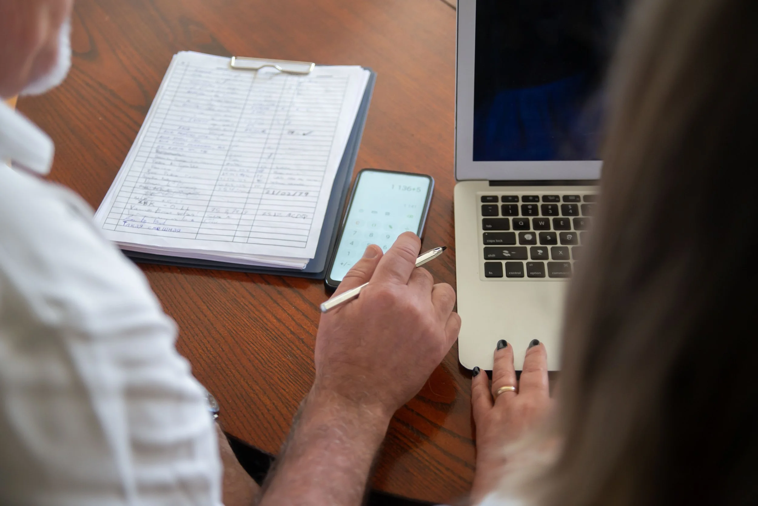 Two people are seated at a wooden table, one is using a smartphone and the other is using a laptop. There is a clipboard with handwritten notes, and the scene appears to be a work or study setting.