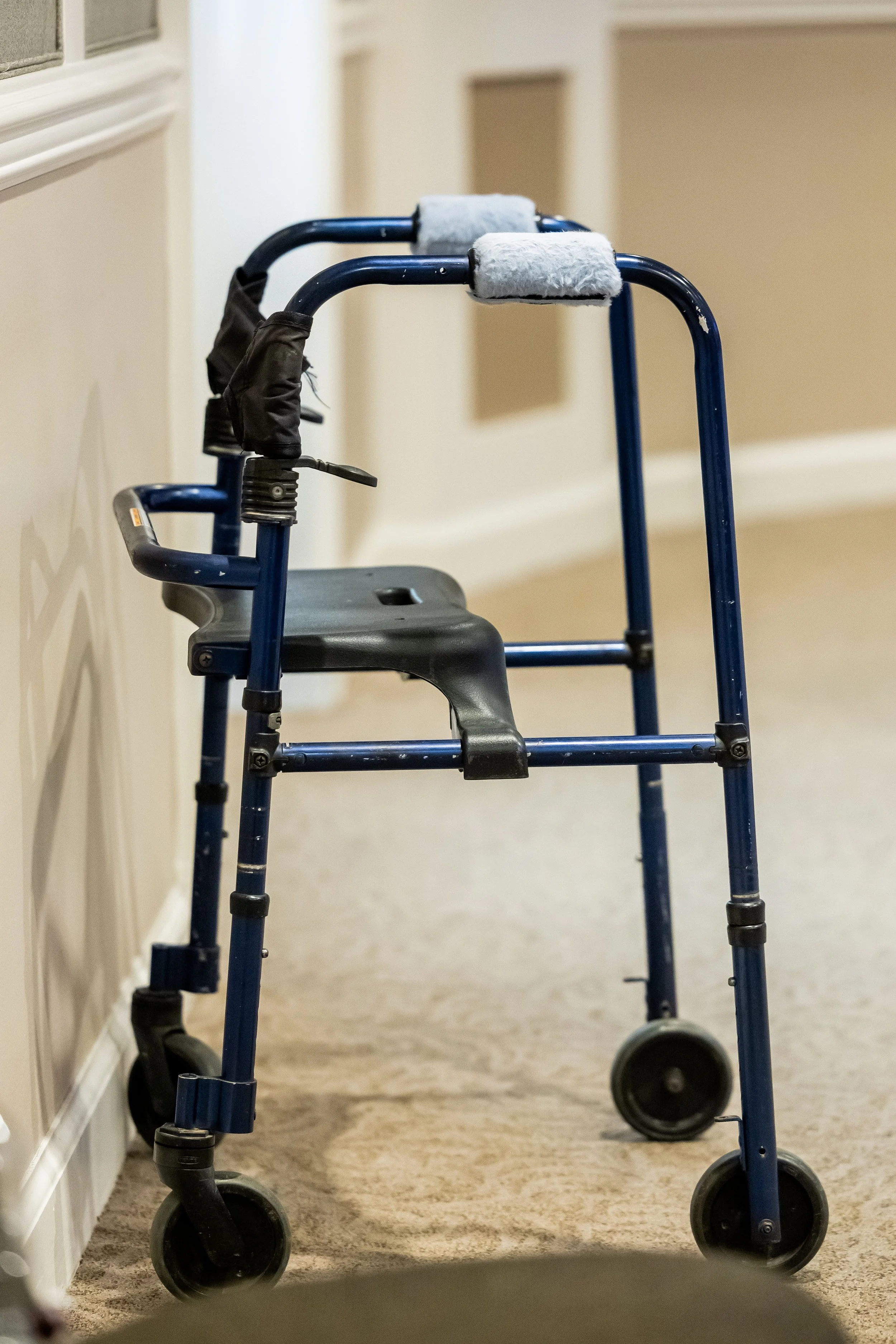 A blue walker with black wheels and padded handgrips, placed in a hallway with beige walls and carpeted floor.