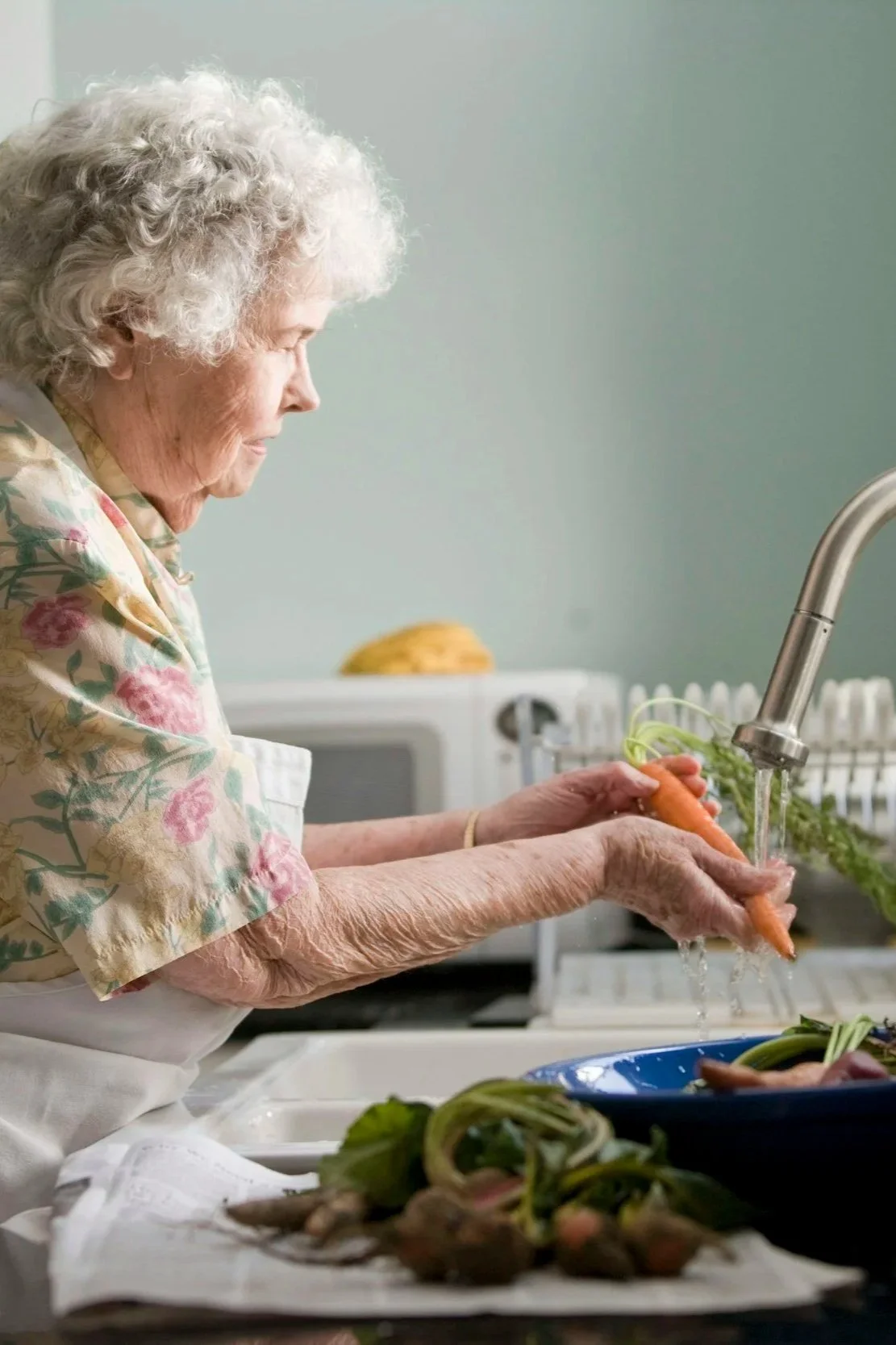 An elderly woman with curly gray hair washing a carrot in a kitchen sink.