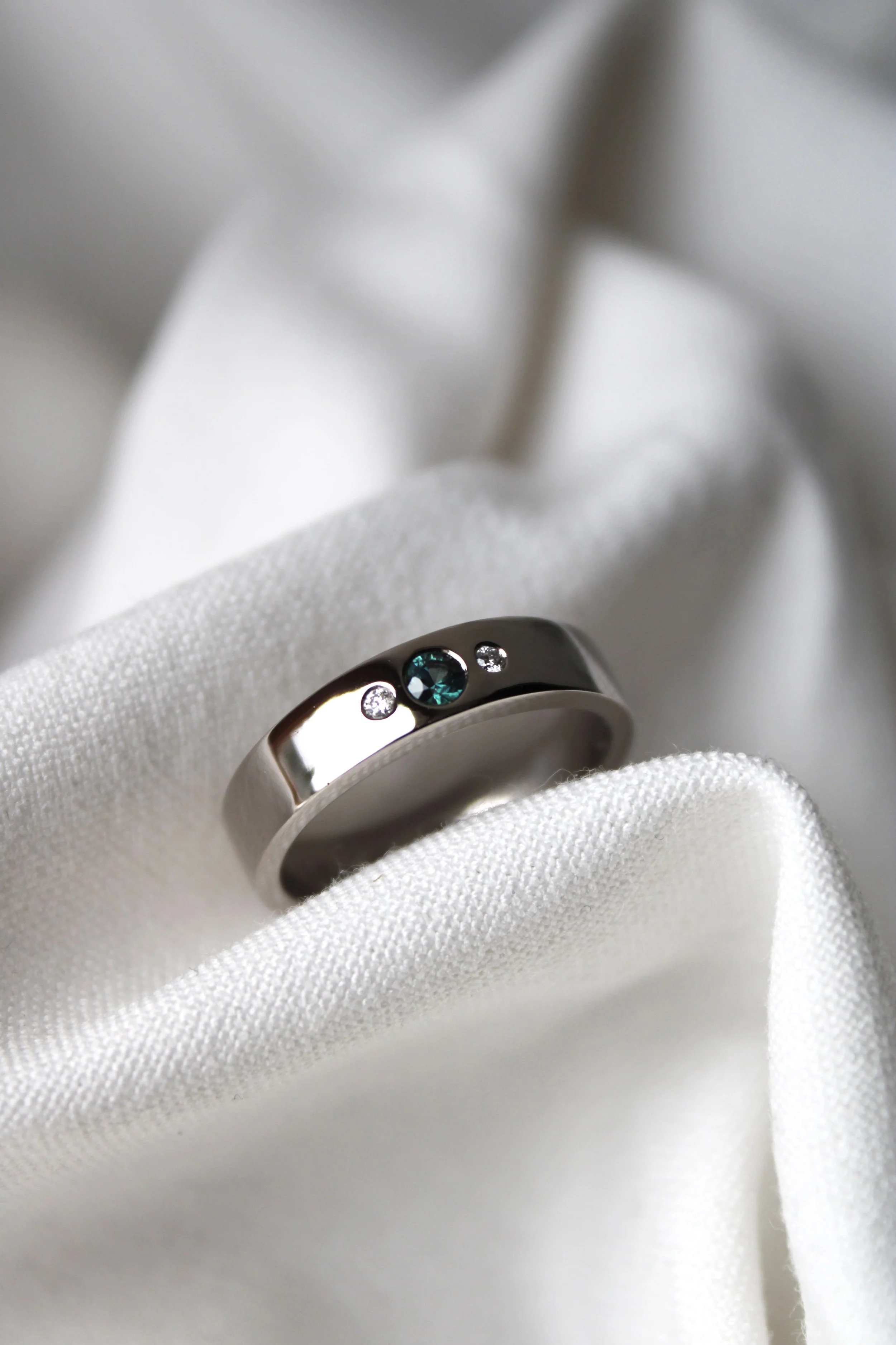 Une bague, alliance, en métal argenté avec une pierre central bleu turquoise et deux petites pierres brillantes, posée sur un tissu blanc.