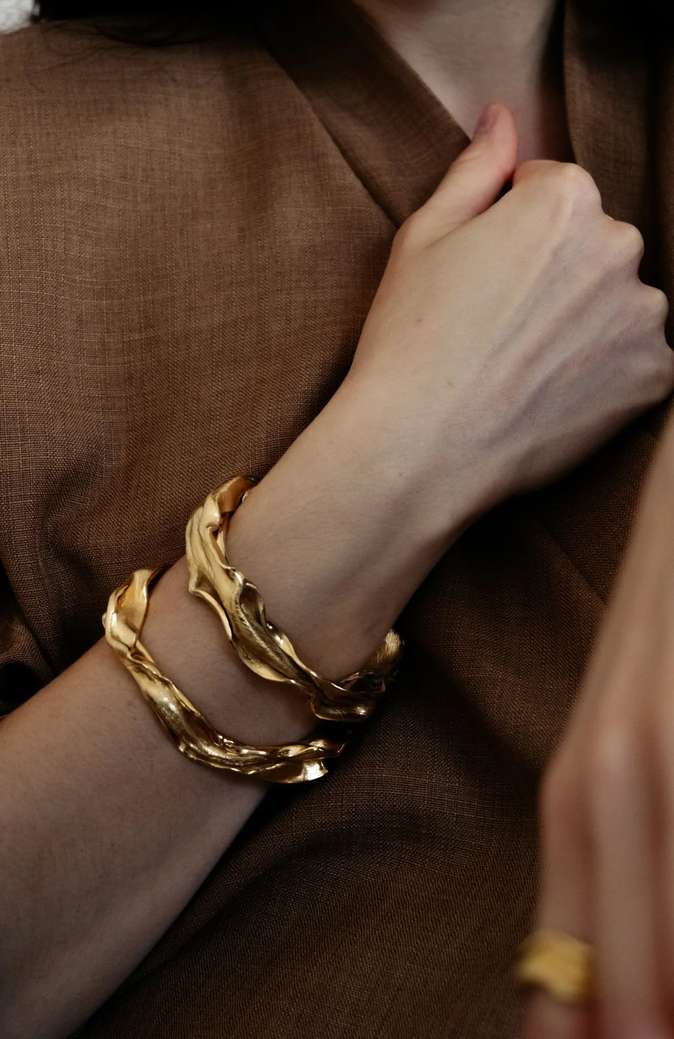 Poignet de femme portant deux bracelets, joncs, avec des formes ondulantes, dorés, vêtue d'une chemise marron.