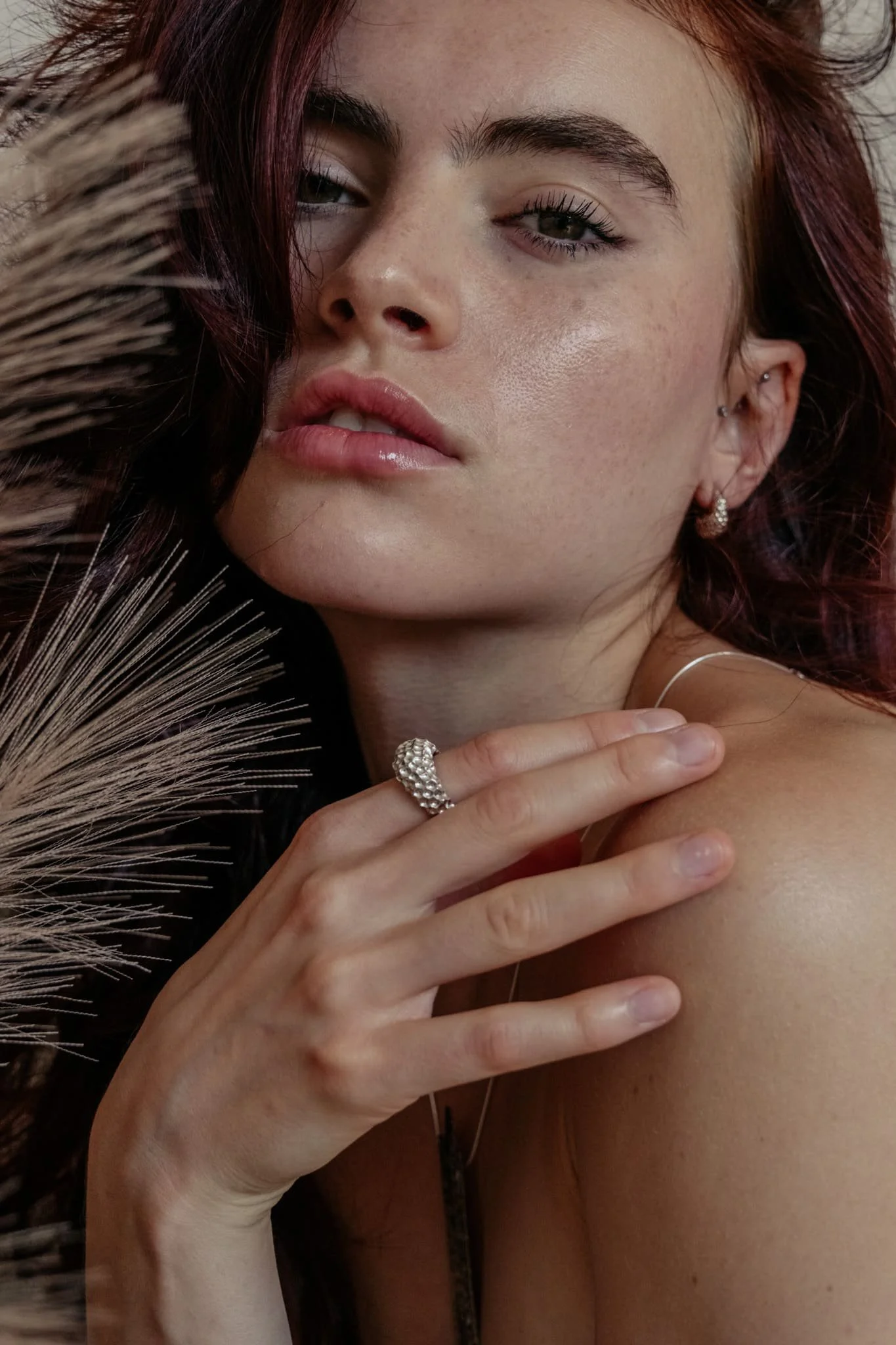 Un portrait rapproché d'une femme avec des cheveux roux, portant des boucles d'oreilles en forme de goutte et une bague argentées martelée, avec une main sur son épaule, regardant vers l'appareil.