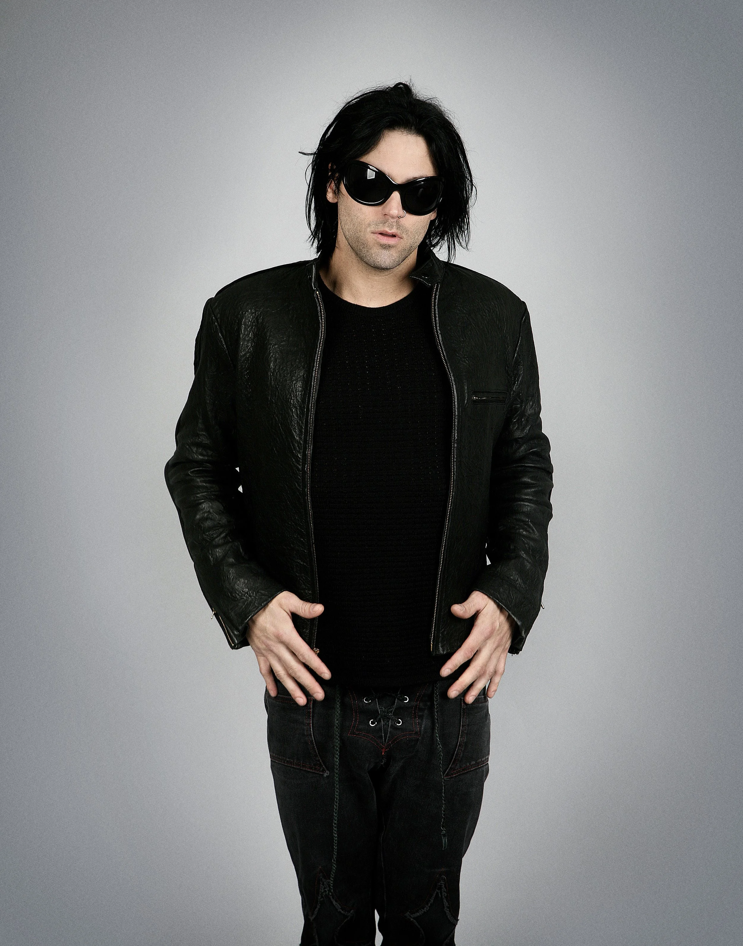 A man with black hair wearing large black sunglasses, a black leather jacket, and black pants, posing against a plain gray background.