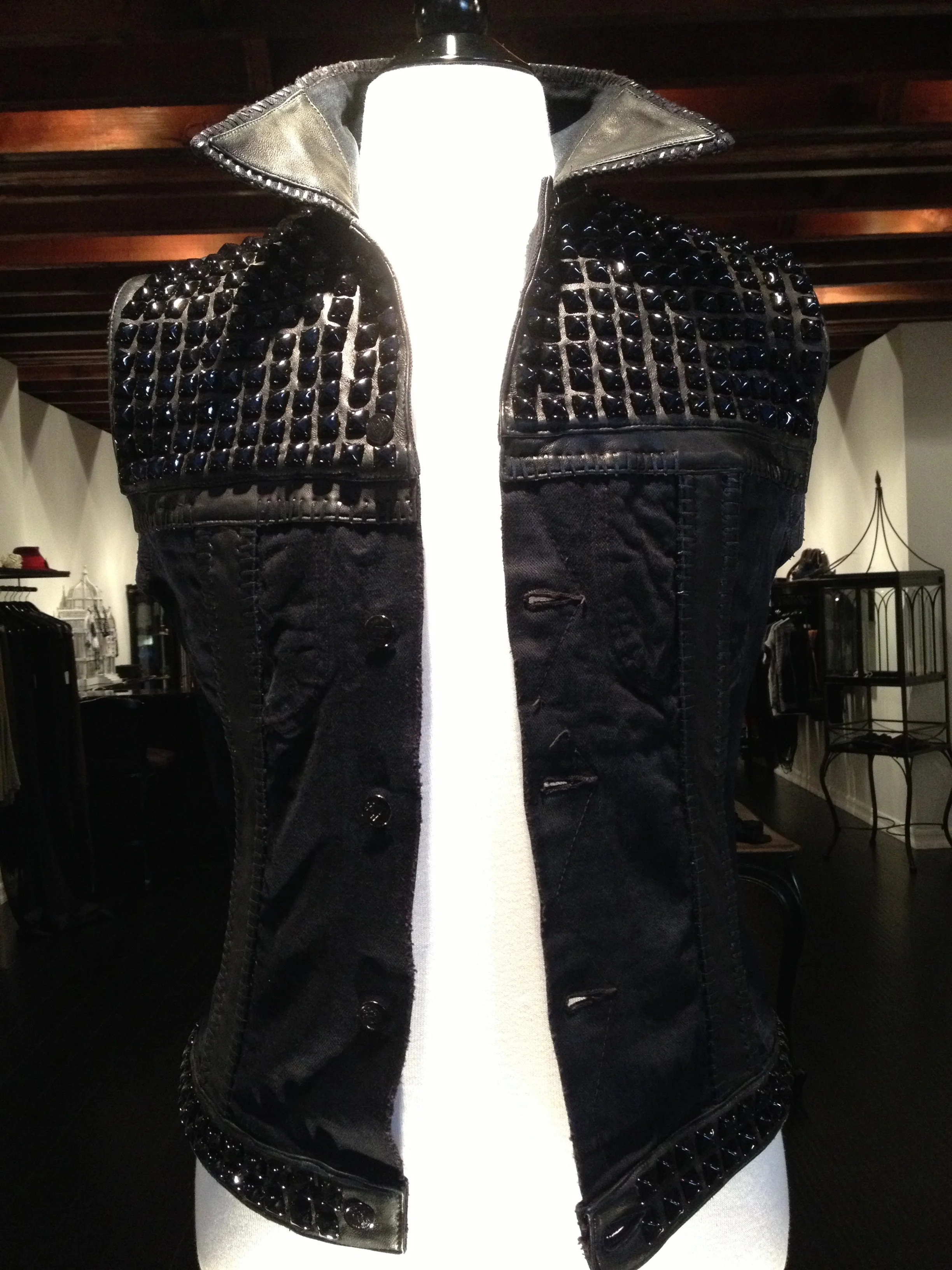 Black denim vest with black studded leather accents on shoulders and hem displayed on a white mannequin.