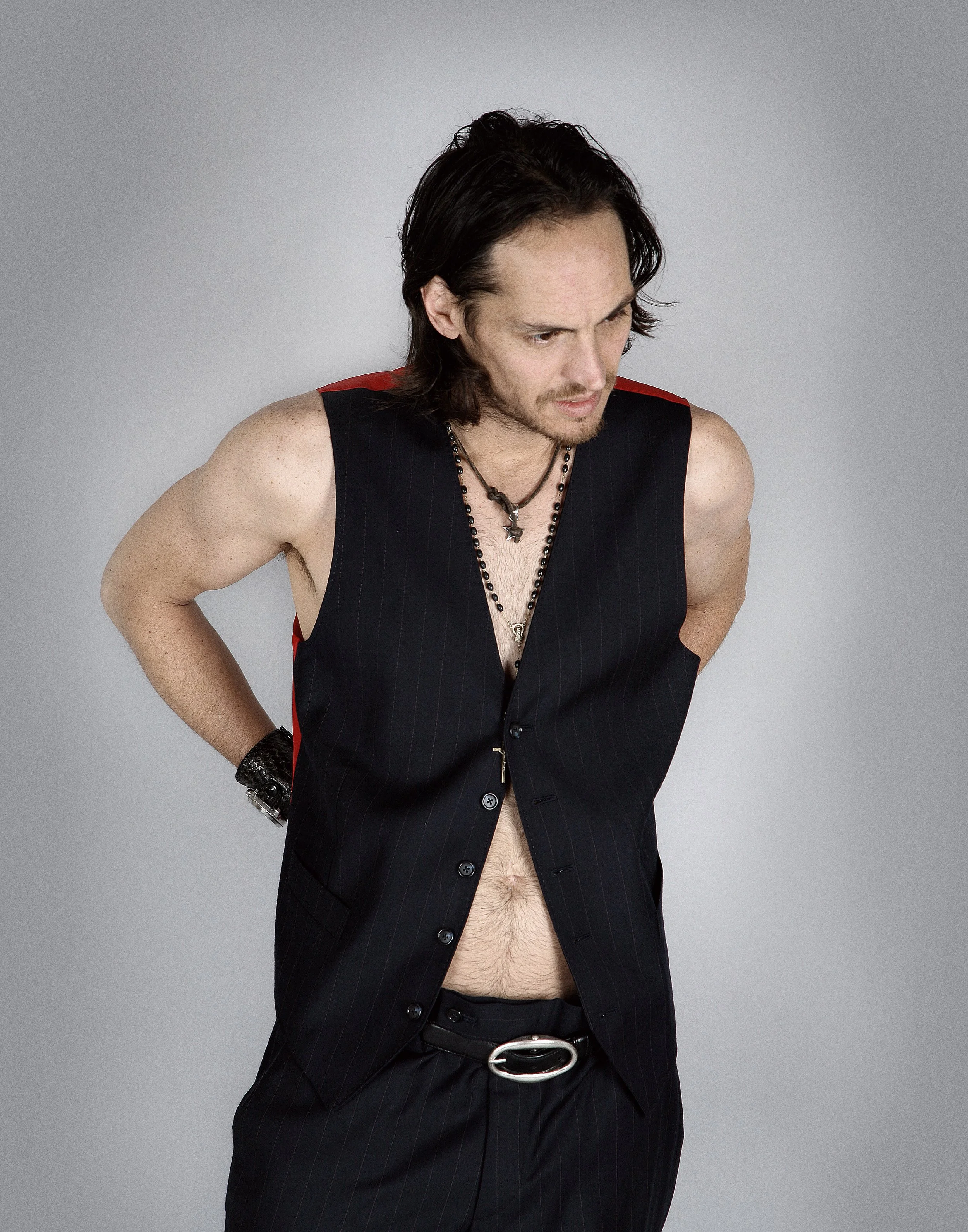 A man with shoulder-length dark hair wearing a sleeveless black pinstripe vest, black pants with a silver belt, and necklaces, looking downward with his hands on his hips against a plain gray background.