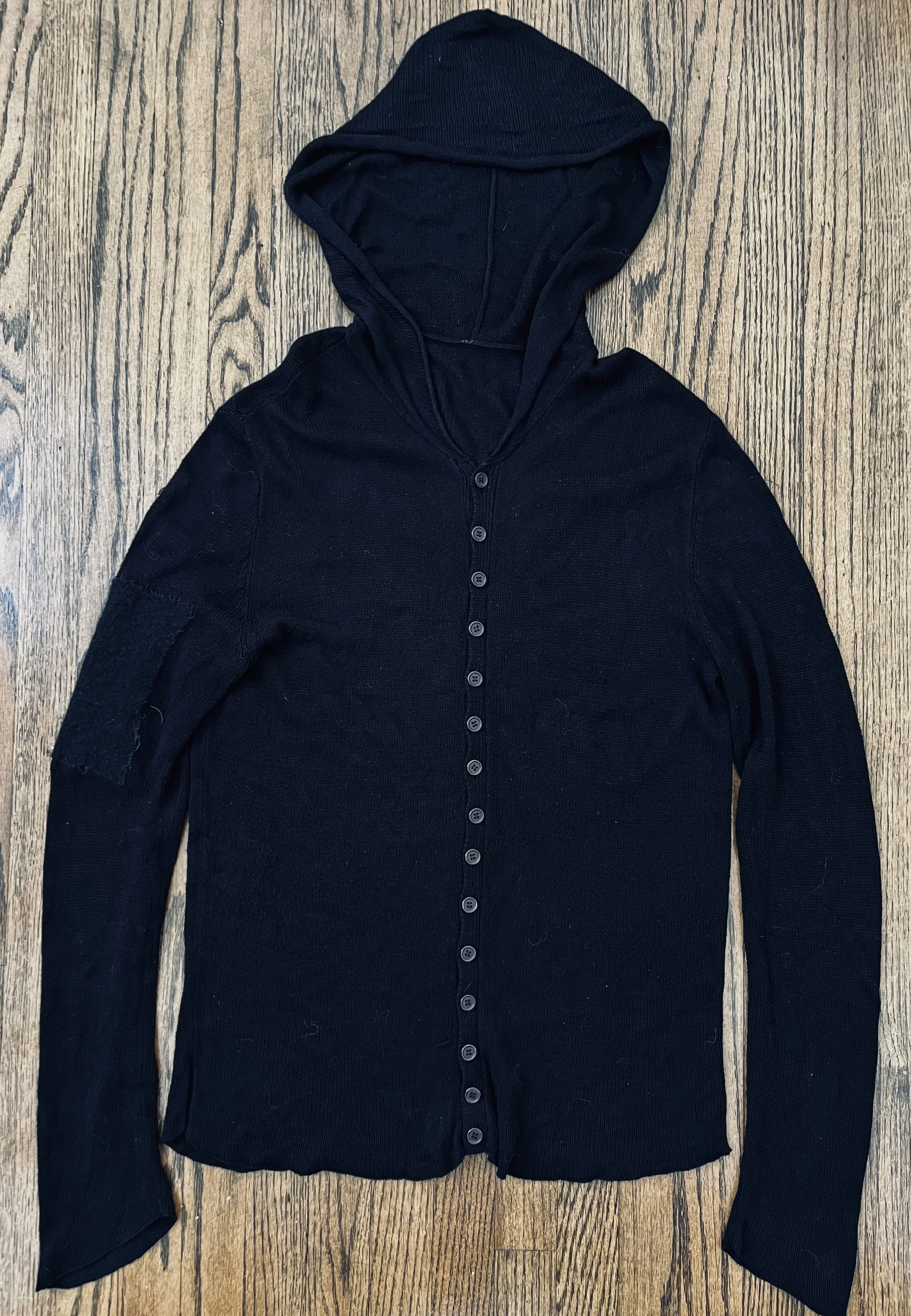 A black hooded cardigan with buttons laid flat on a wooden floor.