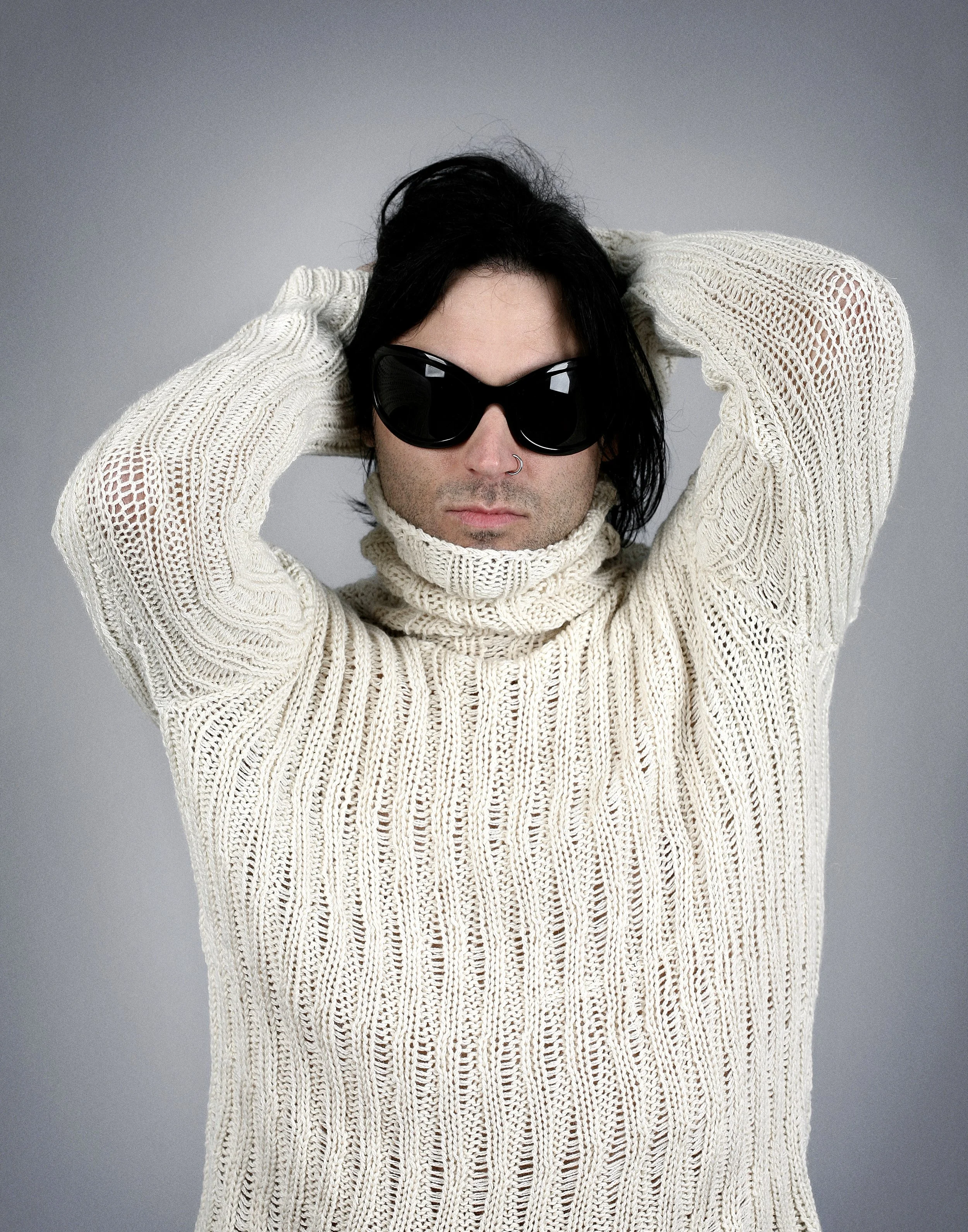 A man with black hair, wearing black sunglasses, a nose ring, and a cream turtleneck sweater, standing against a gray background.