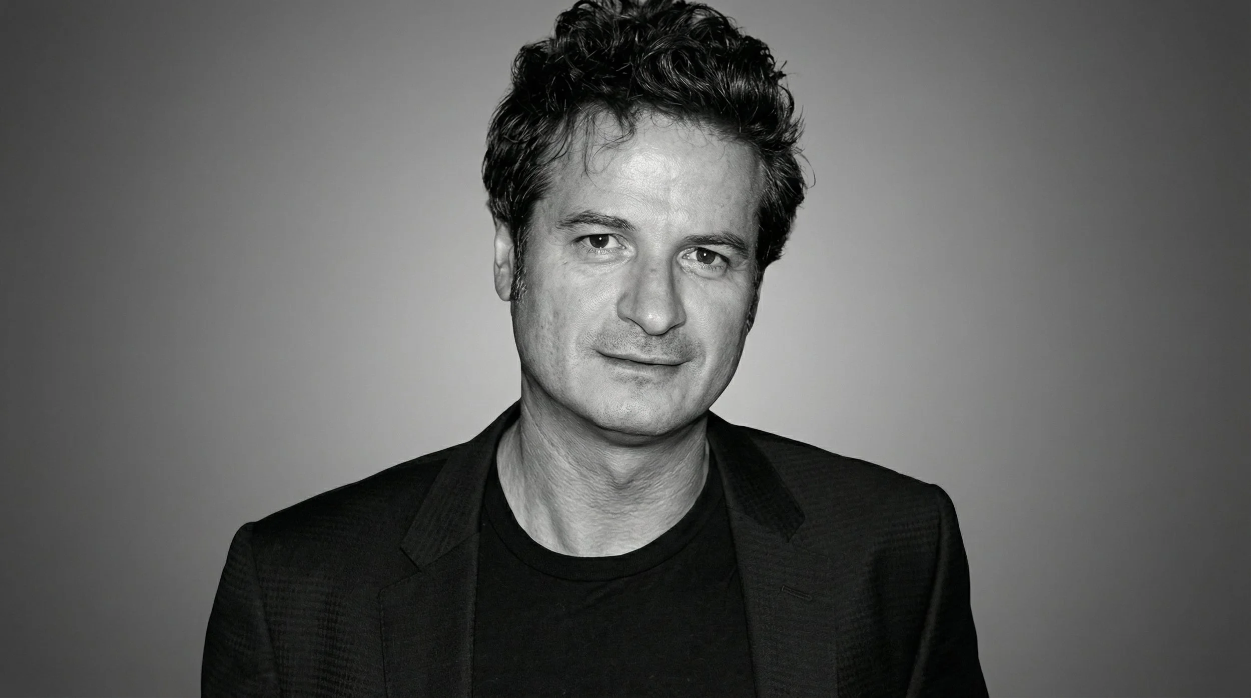 Black and white portrait of a man with wavy hair, wearing a dark blazer over a t-shirt, looking at the camera with a slight smile.