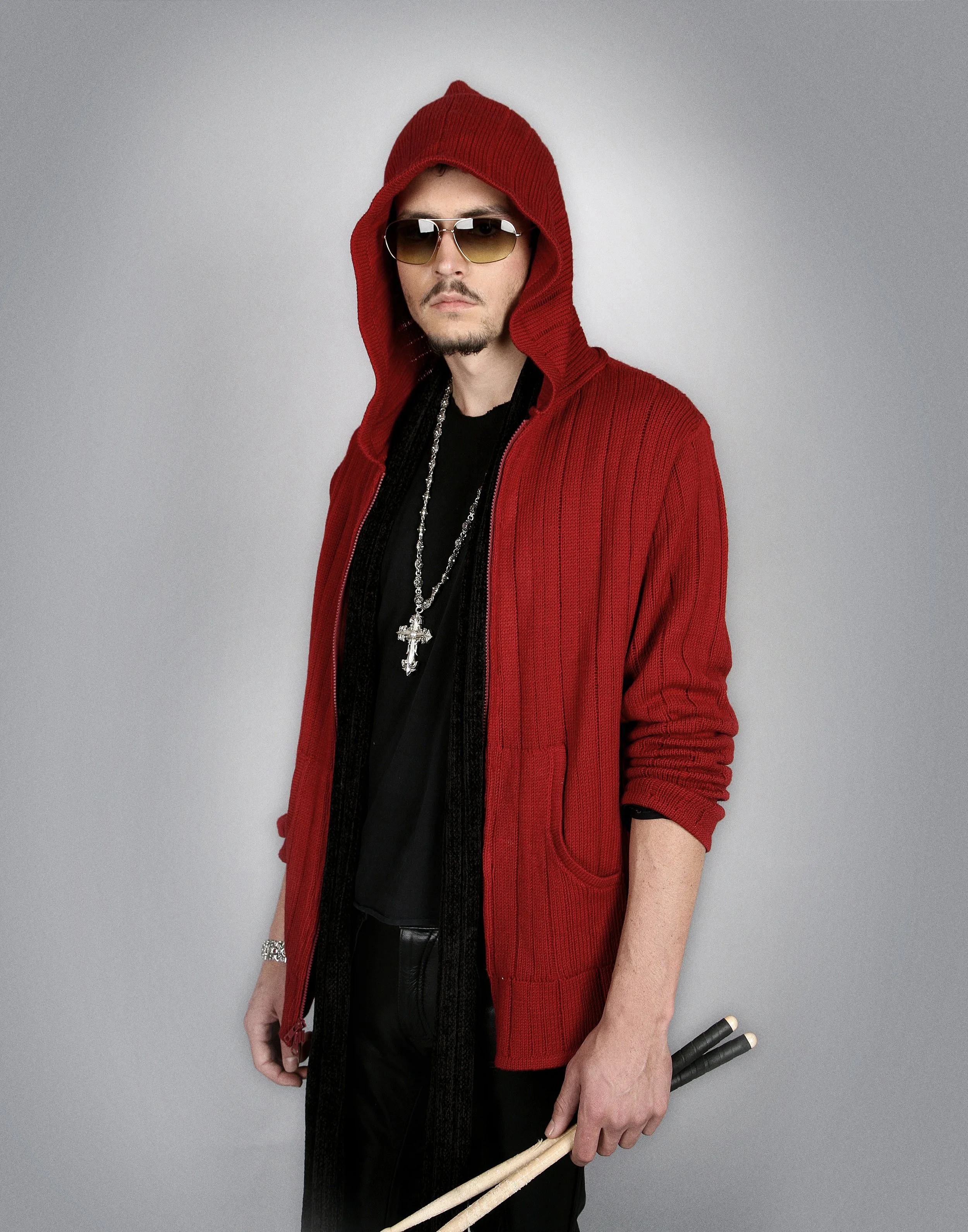 Man dressed in a red hooded sweater, black shirt, sunglasses, with jewelry, holding drumsticks, standing against a gray background.