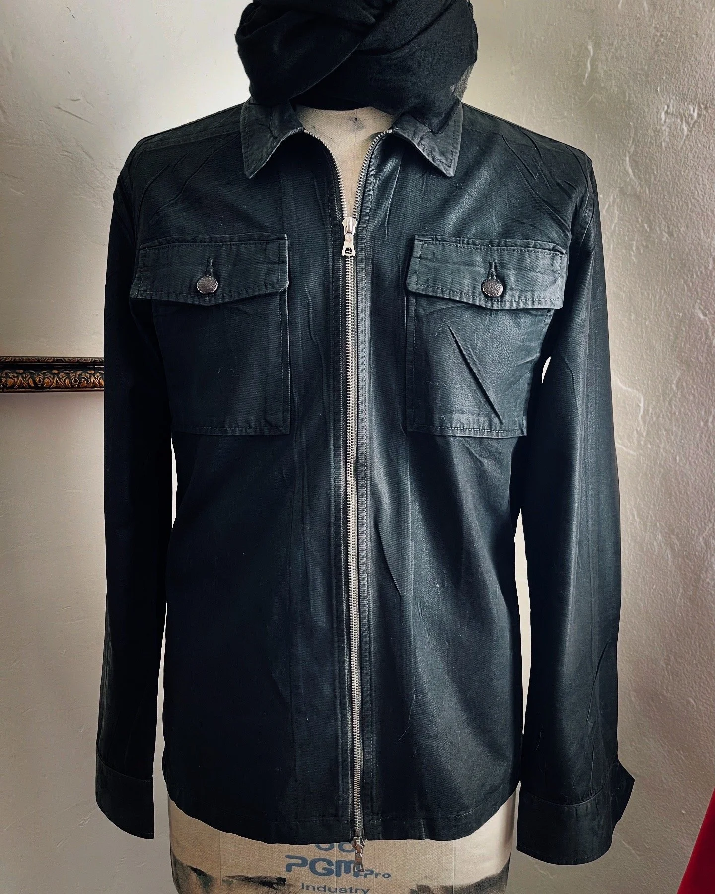 Black leather jacket with two chest pockets and a front zipper on a mannequin with a beige torso and a black cloth covering its neck.