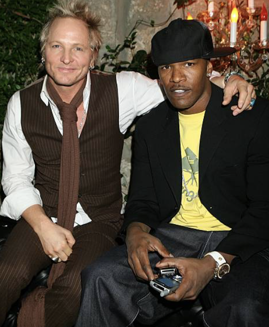 Two men sitting close together at a festive indoor gathering. The man on the left has light skin, blond hair, and is wearing a brown pinstripe vest over a white shirt with rolled-up sleeves, and a brown tie. The man on the right has dark skin, black 