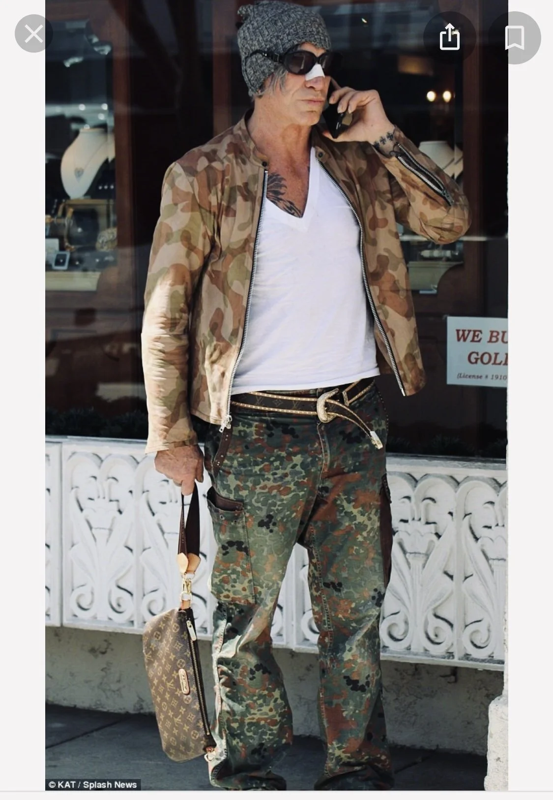 A man standing outside a jewelry store, talking on his cellphone, wearing sunglasses, a gray beanie, a camouflage jacket, white t-shirt, camo cargo pants with brown patches, a gold belt, and holding a Louis Vuitton handbag.