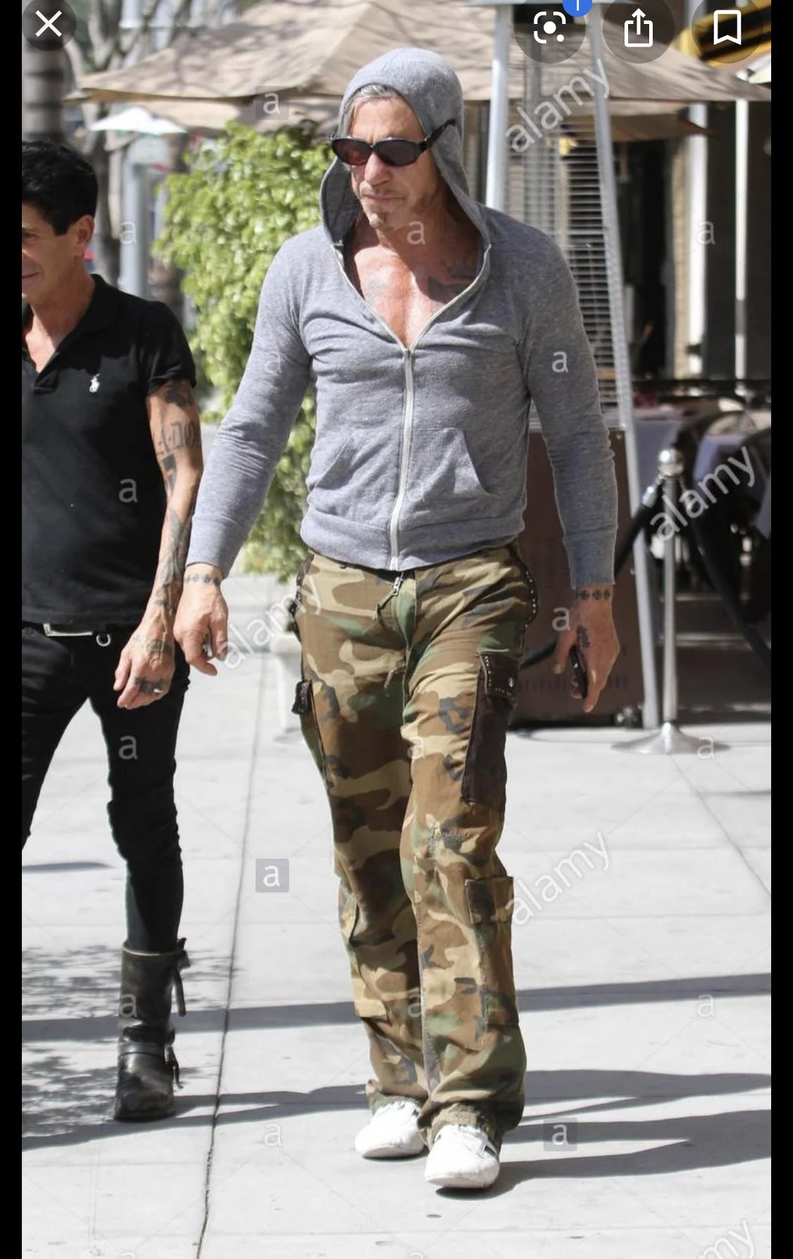 Man wearing a gray zip-up hoodie with the hood up, sunglasses, camouflage cargo pants, and white sneakers, walking outside on a sunny day