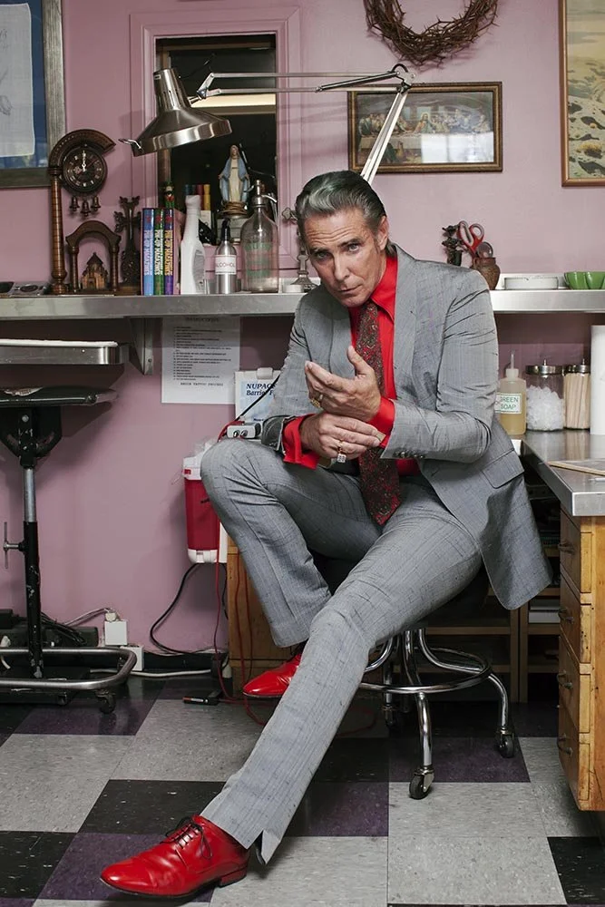 A person in a gray suit with red shirt and red shoes sitting on a stool in a room with pink walls, holding their shoulder and looking at the camera.