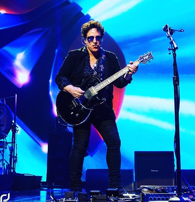 A male guitarist performing on stage, wearing sunglasses, a black jacket, and dark pants, with colorful stage lighting in the background.