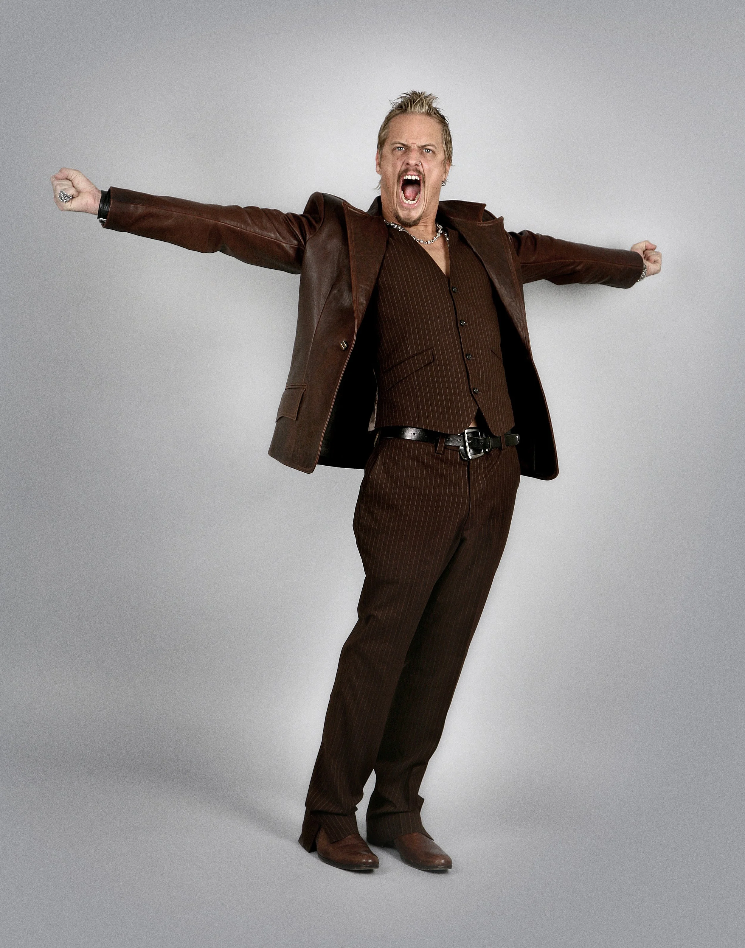 Man with blond hair, wearing a brown pinstripe suit and leather jacket, shouting with arms outstretched against a plain gray background.