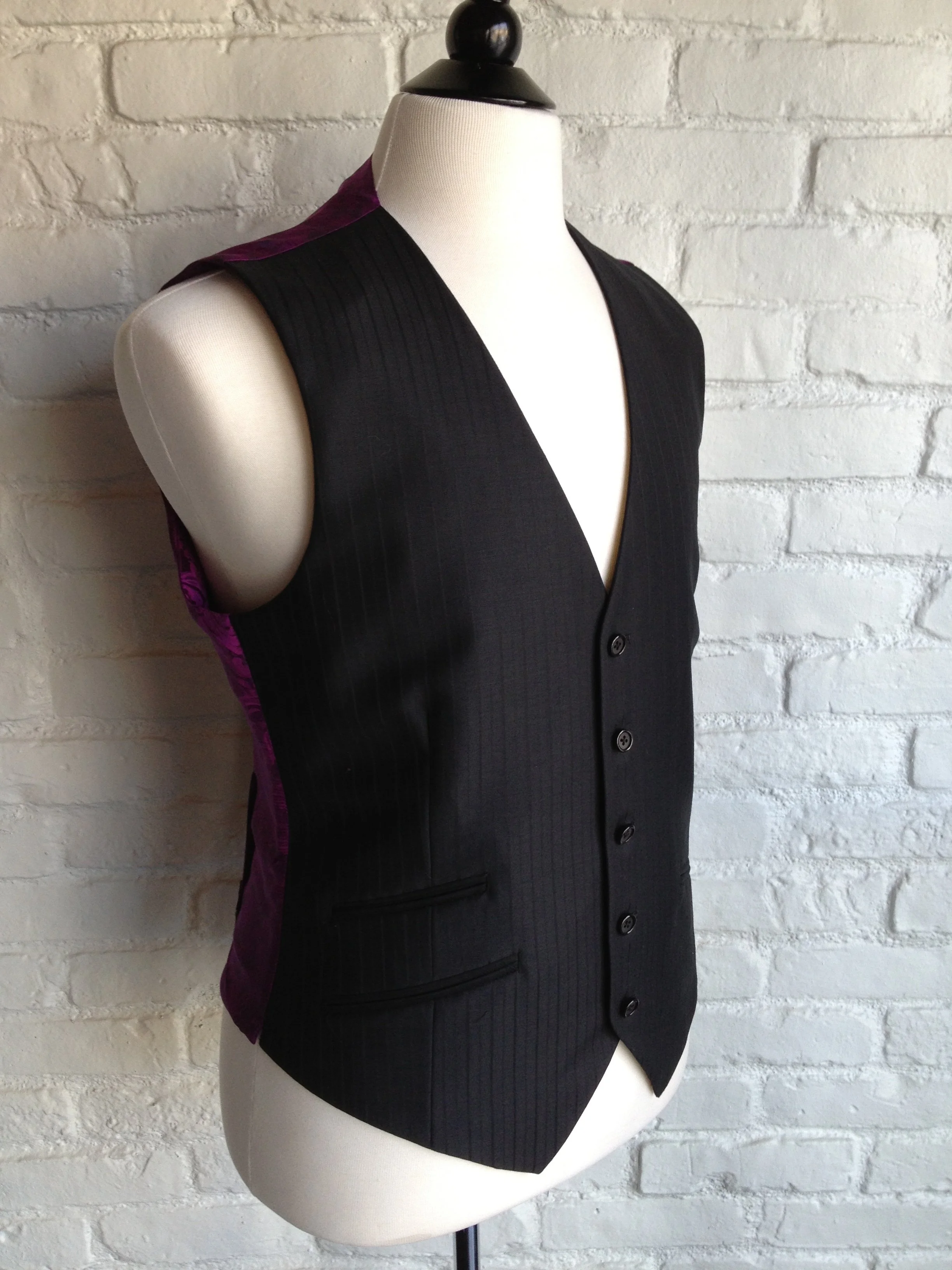 A sleeveless black vest with purple accents on a mannequin, placed against a white brick wall.