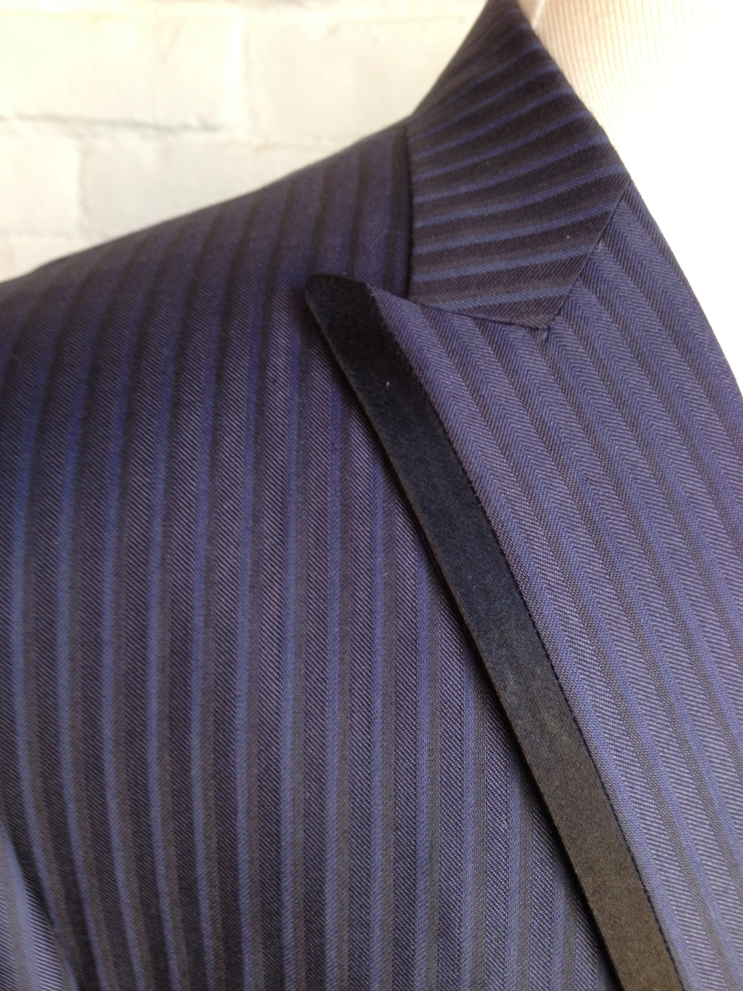 Close-up of a navy blue pinstripe suit jacket with a black velvet lapel on a white mannequin, against a white brick wall background.