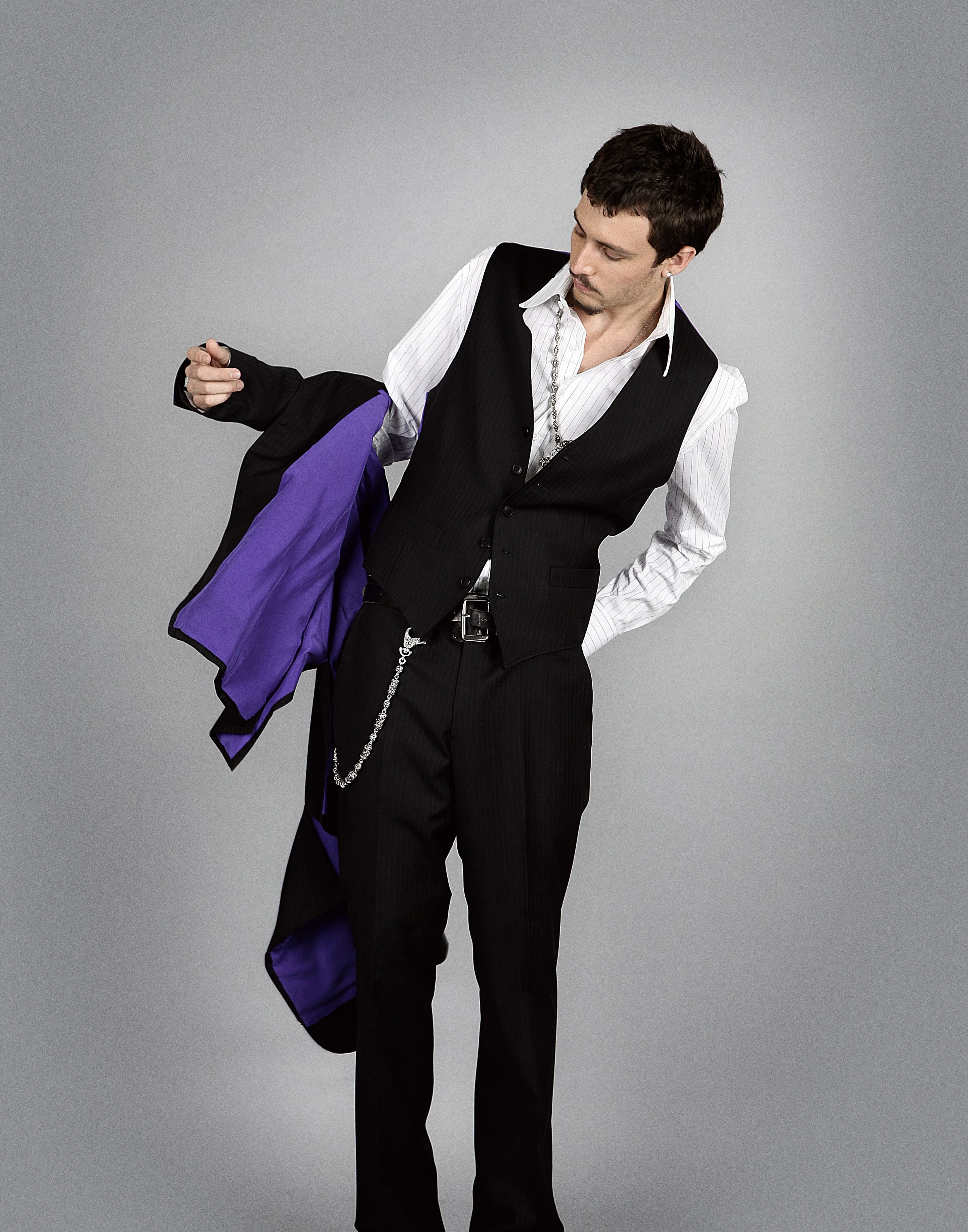 Man in black suit with white shirt and purple lining, looking down, holding jacket over shoulder, posing against a grey background.