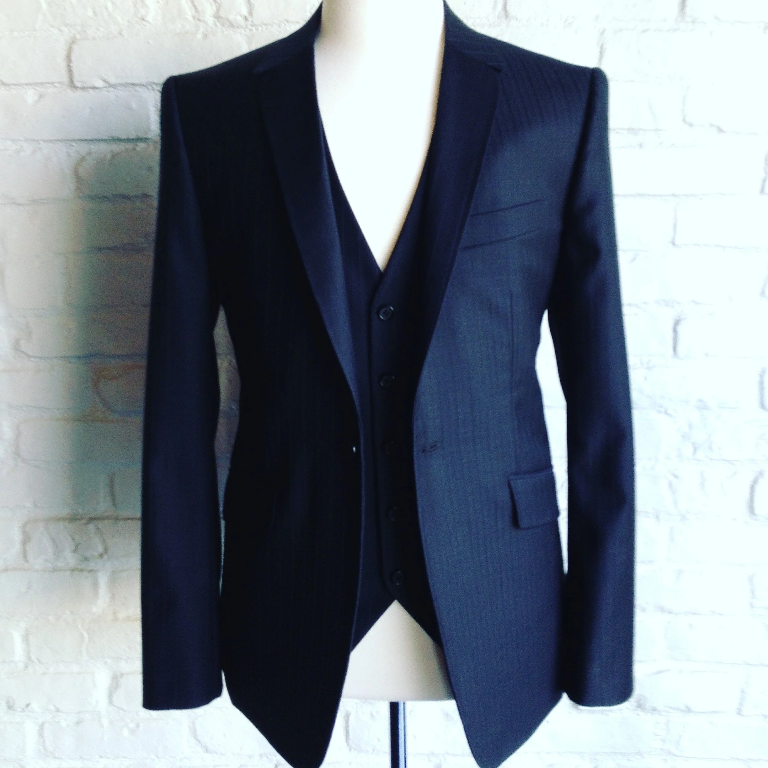A mannequin dressed in a dark navy blue suit with a single-breasted blazer and matching vest, standing against a white brick wall.