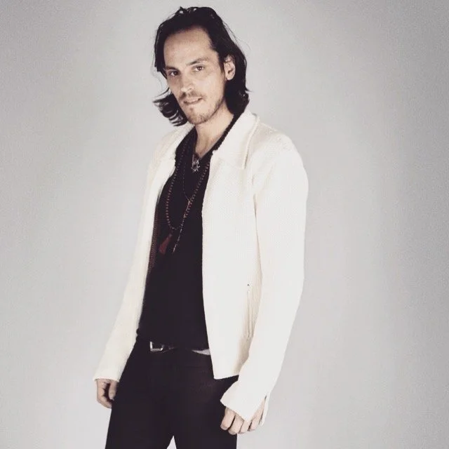 A young man with shoulder-length dark hair and a slight beard, wearing a white jacket over a black shirt and black pants, standing against a plain light gray background.