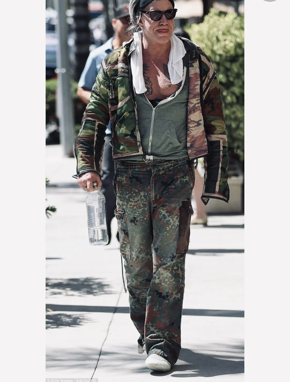 Woman walking outdoors in camouflage clothing, sunglasses, and a camo jacket, holding a water bottle.