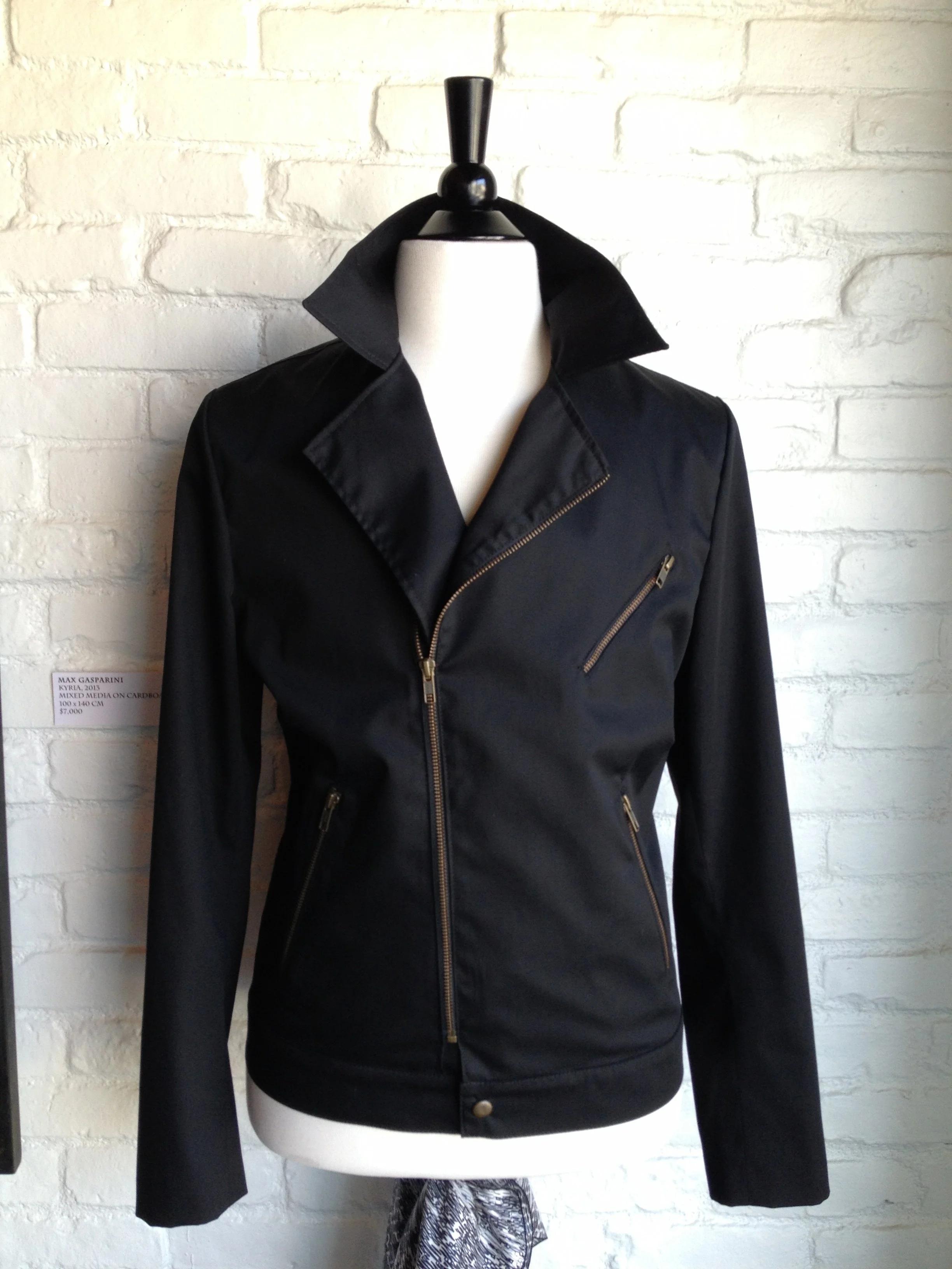 Black leather jacket with gold zippers on a dress form, against a white brick wall.