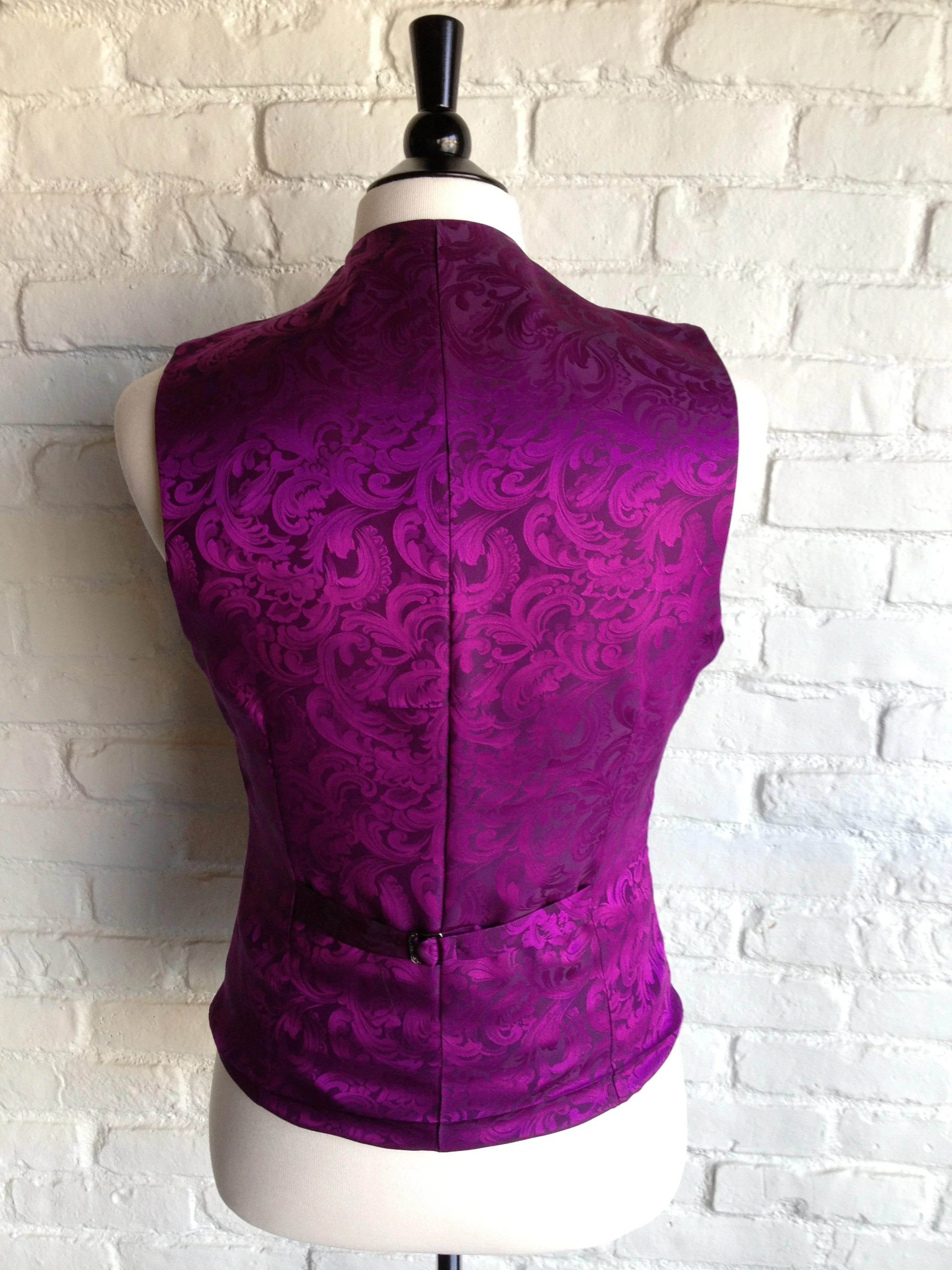 Back view of a sleeveless, purple, brocade vest with a floral pattern on a mannequin against a white brick wall.